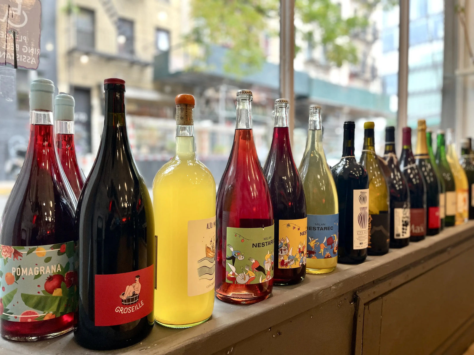 A row of assorted glass wine and sparkling beverage bottles with colorful labels, lined up on a wooden counter in front of a window with a blurred street scene outside.
